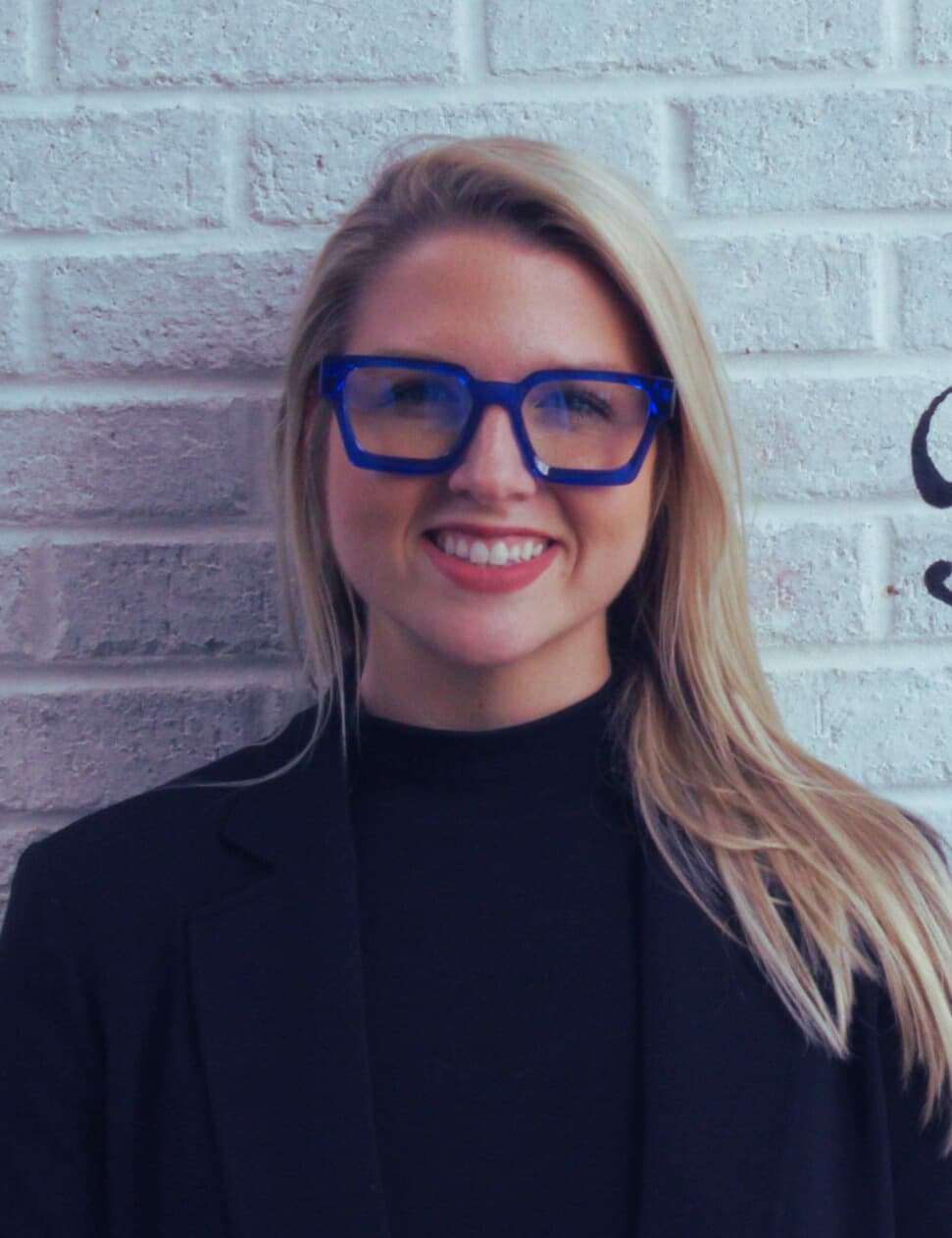 Meghan Celo smiling in her signature cobalt-blue glasses, in front of a white brick wall